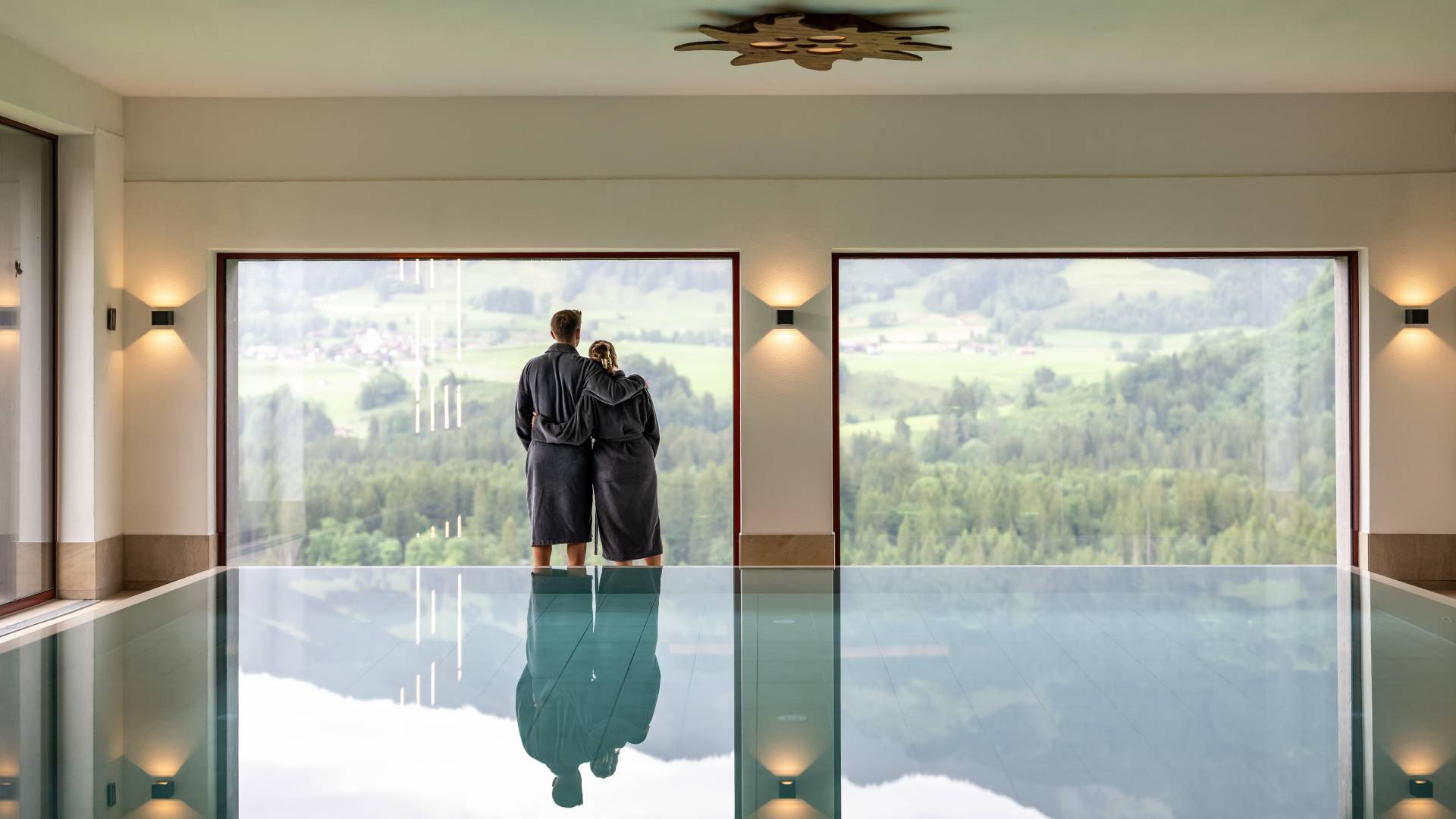 Paar im Bademantel stehen zwischen Pool und Panoramafenster