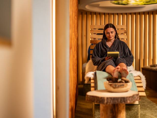 Frau in Bademantel sitz auf einer Holzliege und ließt ein Buch